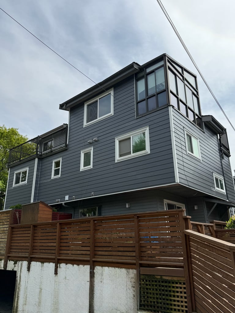 Modern two-story blue-gray house with white trim windows, elevated on stilts, surrounded by wooden fence, under cloudy sky