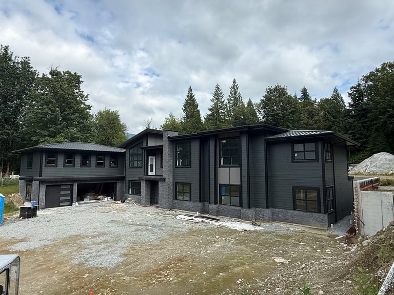 Modern black residential home under construction on gravel lot surrounded by evergreen trees and cloudy sky
