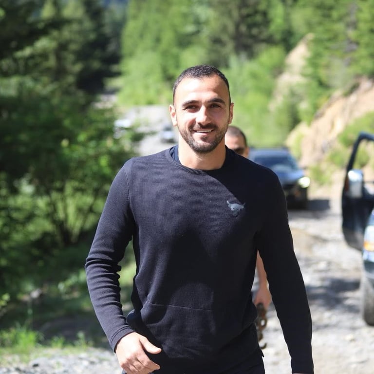 Man with dark hair and beard wearing a black long-sleeved shirt standing on a mountain road with trees and parked car in background